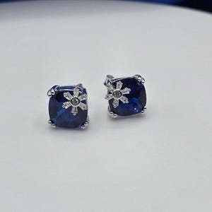 May include: A pair of stud earrings featuring square-cut deep blue stones set in silver-toned metal. Each earring is accented with a small, clear, star-shaped crystal cluster positioned on the surface of the blue stone, secured by metal prongs.
