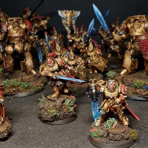 Op de afbeelding: Een verzameling miniatuurfiguren, voornamelijk goud en rood, die gepantserde krijgers met zwaarden en schilden uitbeelden. De figuren zijn gerangschikt op kleine, getextureerde bases, wat wijst op een fantasie- of sciencefictionthema.