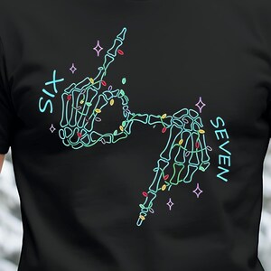 Puede incluir: Camiseta negra con un diseño verde neón y azul de dos manos de esqueleto que forman los números seis y siete. Las manos están decoradas con luces de colores y las palabras "SIX" y "SEVEN" están escritas verticalmente.