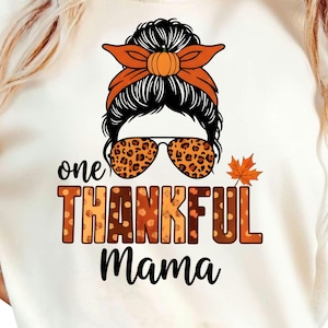 May include: Cream-coloured t-shirt with a graphic featuring a woman's face with a pumpkin-shaped bun, orange headband, and leopard print sunglasses. The text reads "one THANKFUL Mama" in autumnal colours.
