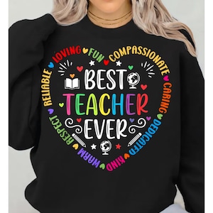 Puede incluir: Sudadera negra con un diseño en forma de corazón. El diseño dice "Best Teacher Ever" en texto colorido, rodeado de palabras como "Loving", "Fun" y "Kind". El diseño incluye estrellas, corazones y un globo terráqueo.
