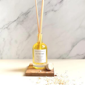 May include: A glass Citrine diffuser bottle with wooden reeds, on a small wooden block. The bottle contains a yellow liquid and has a white label with the text "WRENSPACE CITRINE Mandarin Bergamot Neroli Handcrafted Natural Vegan". A white stopper and crystals are scattered nearby.