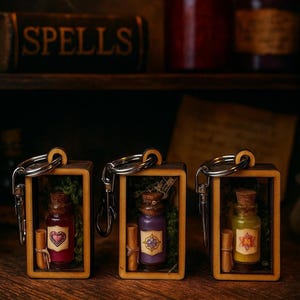May include: Three miniature potion bottle keychains in wooden frames. Each bottle is a different color: red, purple, and yellow-green. The bottles have cork stoppers and are decorated with a heart, a floral design, and a scroll. The word "SPELLS" is visible in the background.