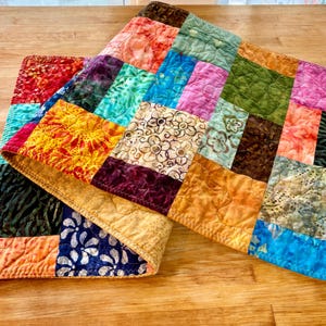 May include: Two colorful patchwork quilts with a variety of patterned squares in shades of red, orange, yellow, green, blue, purple, and brown. The quilts are folded and placed on a wooden surface.