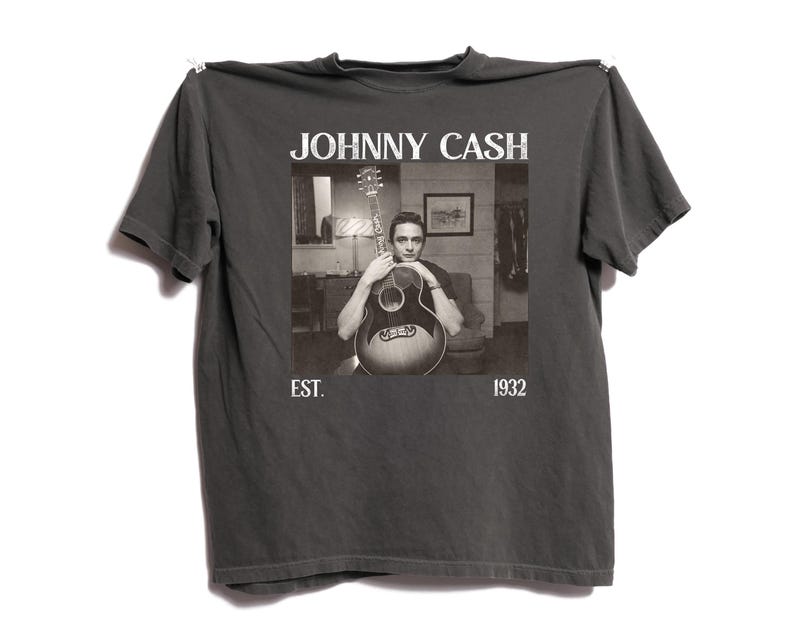 May include: Dark gray t-shirt featuring a black and white image of Johnny Cash holding a guitar. The text "JOHNNY CASH" is above the image, with "EST. 1932" below. The shirt is hanging against a white background.
