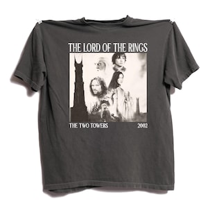 Può includere: T-shirt grigio scuro con un'immagine in bianco e nero del film "Il Signore degli Anelli: Le Due Torri". La grafica include il titolo del film, ritratti di personaggi e l'anno 2002.