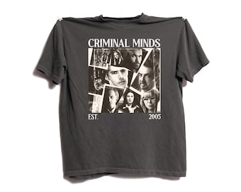 May include: Dark gray t-shirt featuring a collage of black and white images of people. The shirt has the text "CRIMINAL MINDS" at the top and "EST. 2005" at the bottom. The shirt is hanging against a white background.