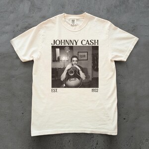 May include: Cream-colored t-shirt featuring a black and white image of Johnny Cash holding a guitar. The text "JOHNNY CASH" is above the image, with "EST. 1932" below. The shirt has a classic crew neck and short sleeves.