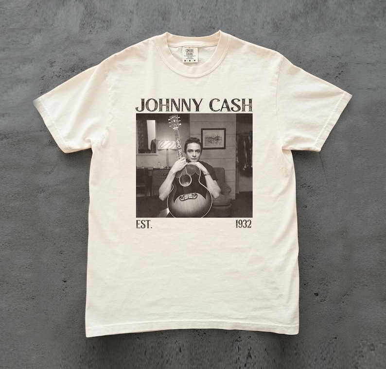 May include: Cream-colored t-shirt featuring a black and white image of Johnny Cash holding a guitar. The text "JOHNNY CASH" is above the image, with "EST. 1932" below. The shirt has a classic crew neck and short sleeves.