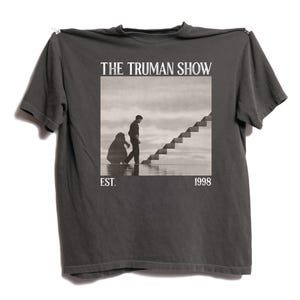 May include: Dark grey t-shirt with a black and white image from "The Truman Show." The image depicts a man ascending stairs with another figure. The text "THE TRUMAN SHOW" is above the image, with "EST. 1998" below.