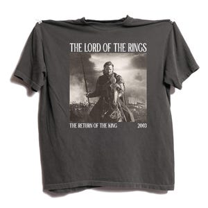 Può includere: T-shirt grigio scuro con un'immagine in bianco e nero de "Il Signore degli Anelli: Il Ritorno del Re". Il testo "THE LORD OF THE RINGS" è sopra l'immagine, con "THE RETURN OF THE KING" e "2003" sotto.