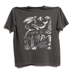 May include: Dark gray t-shirt featuring a white illustration of various sea creatures. The design includes a sea turtle, whale, dolphin, octopus, seahorse, and several types of fish and aquatic plants. The shirt is hanging against a white background.
