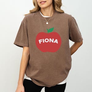 May include: Brown t-shirt with a large red apple graphic. The apple has a green leaf and the name "FIONA" in white letters. The shirt has short sleeves and a crew neck.
