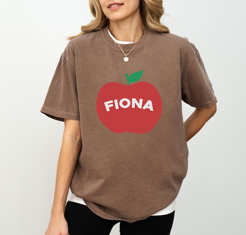 May include: Brown t-shirt with a large red apple graphic. The apple has a green leaf and the name "FIONA" in white letters. The shirt has short sleeves and a crew neck.