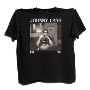 May include: Black t-shirt featuring a black and white image of Johnny Cash holding a guitar. The text "JOHNNY CASH" is above the image, with "EST. 1932" below. The shirt is hanging against a white background.