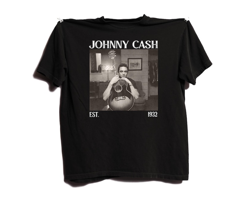 May include: Black t-shirt featuring a black and white image of Johnny Cash holding a guitar. The text "JOHNNY CASH" is above the image, with "EST. 1932" below. The shirt is hanging against a white background.