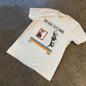 May include: Off-white t-shirt with the text "THE GIRL NEXT DOOR" in black. The shirt features a graphic of a woman and a man in a square, with the text "EST. 2004" below a striped design in orange, blue, and yellow.