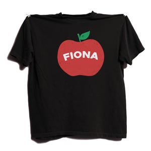 May include: Black t-shirt featuring a red apple graphic with a green leaf and the name "FIONA" in white letters. The shirt is hanging on a white background.
