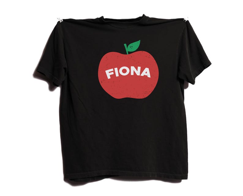 May include: Black t-shirt featuring a red apple graphic with a green leaf and the name "FIONA" in white letters. The shirt is hanging on a white background.