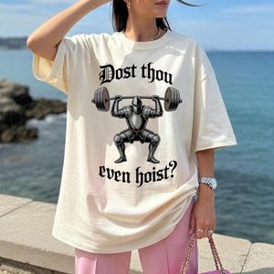 May include: Cream-colored t-shirt featuring a black graphic of a knight lifting a barbell, with the text "Dost thou even hoist?" in a gothic font. The shirt has short sleeves and a relaxed fit.