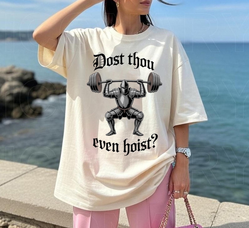 May include: Cream-colored t-shirt featuring a black graphic of a knight lifting a barbell, with the text "Dost thou even hoist?" in a gothic font. The shirt has short sleeves and a relaxed fit.
