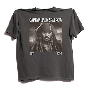 Pode incluir: Camiseta cinza escuro com uma imagem em preto e branco do Capitão Jack Sparrow. O texto "CAPTAIN JACK SPARROW" está acima da imagem, com "EST. 1690" abaixo. A camiseta está pendurada em um fundo branco.