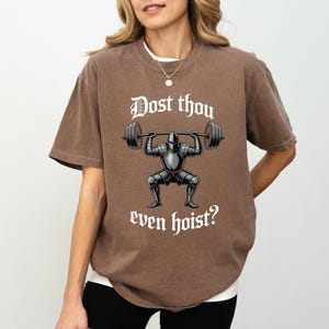 May include: Brown t-shirt with the text "Dost thou even hoist?" above an illustration of a knight lifting weights. The knight is wearing armor and the barbell has weights on each end. The shirt has short sleeves.