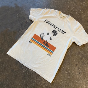 May include: Off-white t-shirt featuring the text "FORREST GUMP" above a graphic of a running figure. The graphic includes a rectangular design with orange, blue, and yellow stripes. The text "EST. 1994" is below the graphic.