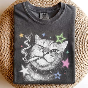 May include: A charcoal gray t-shirt featuring a white graphic of a winking cat wearing a spiked collar and smoking a cigarette. The design includes colorful stars in yellow, green, pink, and blue. The inner neck label reads Comfort Colors.