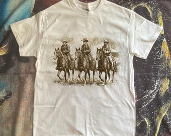 May include: White t-shirt featuring a sepia-toned graphic of three cowboys riding horses. The cowboys wear hats and jackets, and the horses are in motion. The shirt has short sleeves and a crew neck.
