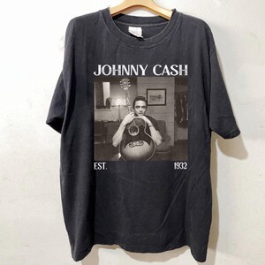 May include: Black t-shirt featuring a black and white image of Johnny Cash holding a guitar. The text "JOHNNY CASH" is above the image, with "EST. 1932" below. The shirt is hanging on a wooden hanger.