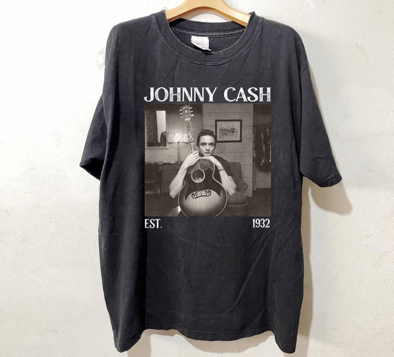 May include: Black t-shirt featuring a black and white image of Johnny Cash holding a guitar. The text "JOHNNY CASH" is above the image, with "EST. 1932" below. The shirt is hanging on a wooden hanger.
