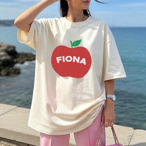 May include: Cream-colored t-shirt featuring a large red apple graphic with the name "FIONA" in white letters. The apple has a green leaf. The shirt has short sleeves and a relaxed fit. The background shows a body of water.