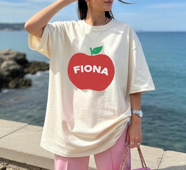 May include: Cream-colored t-shirt featuring a large red apple graphic with the name "FIONA" in white letters. The apple has a green leaf. The shirt has short sleeves and a relaxed fit. The background shows a body of water.