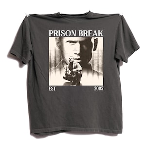 May include: Dark gray t-shirt featuring a graphic from the TV show "Prison Break." The design includes the show's title, a sepia-toned image of a man, and the text "EST. 2005."