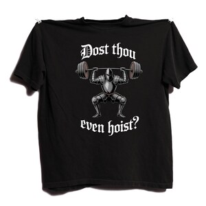 May include: Black t-shirt featuring a graphic of a knight lifting weights, with the text "Dost thou even hoist?" in white. The knight is in silver armor, and the barbell has dark gray weights. The shirt is hanging against a white background.