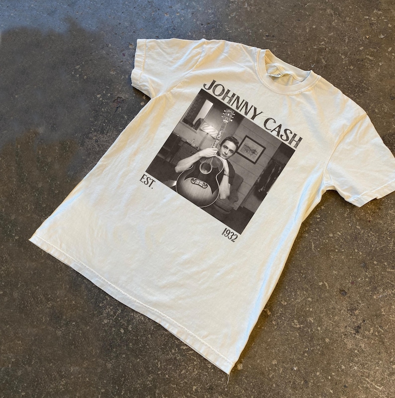 May include: Off-white t-shirt featuring a black and white image of Johnny Cash holding a guitar. The text "JOHNNY CASH" is above the image, with "EST. 1932" below. The shirt has short sleeves and a crew neck.
