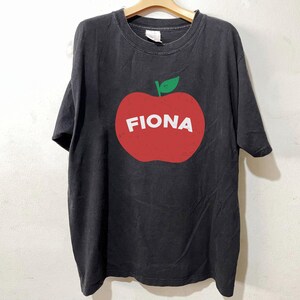 May include: Black t-shirt with a large red apple graphic. The apple has a green leaf and the name "FIONA" in white block letters. The shirt is hanging on a wooden hanger.