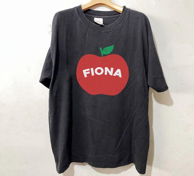 May include: Black t-shirt with a large red apple graphic. The apple has a green leaf and the name "FIONA" in white block letters. The shirt is hanging on a wooden hanger.