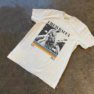 Op de afbeelding: Wit t-shirt met een afbeelding van Chewbacca. De tekst "CHEWBACCA" staat boven de afbeelding en "EST. 1978" eronder. Het ontwerp heeft een retro-stijl met een zwarte achtergrond en kleurrijke strepen.
