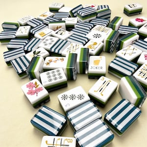 May include: A collection of rectangular Mahjong tiles featuring white faces with various black, green, and pink illustrations, including floral patterns, numbers, and a Joker tile. The backs of the tiles display a striped pattern in dark green and white.