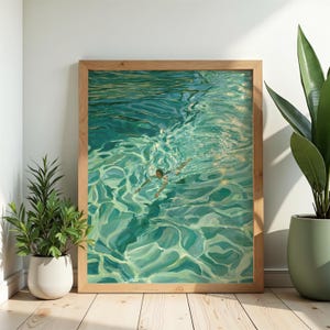 Könnte beinhalten: Ein gerahmter Kunstdruck, der eine schwimmende Person in türkisfarbenem Wasser zeigt. Das Kunstwerk besticht durch lebendige Türkis-, Grün- und Hellblautöne und fängt die wellige Wasseroberfläche mit Sonnenlichtreflexionen und einem Schwimmer ein.