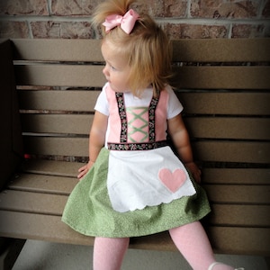 May include: A young child wearing a traditional pink and green dress with a white apron and a pink heart. The dress has floral embroidery and a black belt. The child is also wearing pink tights, white shoes, and a pink bow in their hair. The text "Ada's Accessories" is visible.