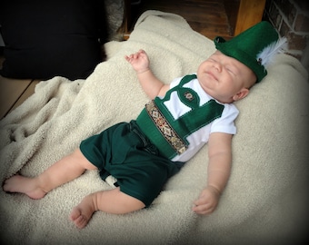 Puede incluir: Un bebé con un traje de lederhosen verde y blanco con un sombrero verde y una pluma blanca. El bebé está acostado sobre una manta blanca.