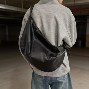 Könnte beinhalten: Eine große, schwarze, lässige Hobo-Umhängetasche mit glatter Textur und breitem Riemen. Die Tasche hat eine geschwungene, halbmondartige Form, die über dem Rücken hängt, mit einem kleinen geknoteten Detail an der Seite, wo der Riemen am Körper ansetzt.