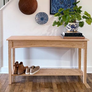 May include: A light-colored wooden console table with a lower shelf and a top surface. The table has a natural wood finish. A pair of brown boots and a pair of beige slip-on shoes are on the lower shelf. A potted plant sits on top.