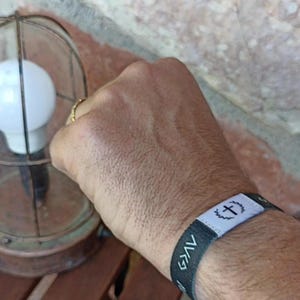 May include: A black and white wristband with a cross symbol and the word "GAV" is worn on a wrist. A vintage-style lamp with a glass enclosure and a light bulb is in the background.