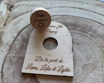 May include: A wooden gift tag and stopper set. The stopper is round with the words "Papy d'amour" and a heart. The tag has a hole and the words "Joyeuse fête Papy" and "De la part de Lana, Lola et Lydie" are engraved on it.