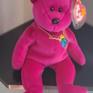 Könnte beinhalten: Ein leuchtend fuchsiafarbener Plüschbär mit einer goldfarbenen Kette, die den Schriftzug 2000 und einen kleinen Globus-Anhänger trägt. Der Bär hat schwarze Kunststoffaugen und eine schwarze Nase sowie ein herzförmiges Ty-Etikett am linken Ohr.