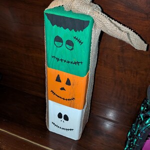 Puede incluir: Una decoración de bloque de madera vertical pintada con tres caras apiladas: un monstruo verde con cabello negro y puntos de sutura, una calabaza naranja y un fantasma blanco. Una cinta de arpillera color canela está atada en la parte superior.